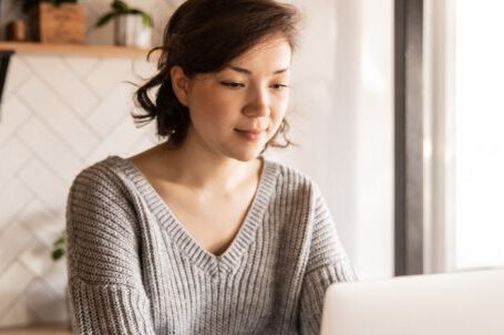 First-time Entrepreneur - Young female in gray sweater sitting at wooden desk with laptop and bottle of milk near white bowl while browsing internet on laptop during free time at home