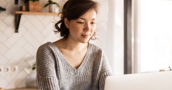 First-time Entrepreneur - Young female in gray sweater sitting at wooden desk with laptop and bottle of milk near white bowl while browsing internet on laptop during free time at home